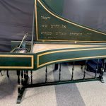 Image of a green harpsichord with open lid. The case and lid are accented with gold boxes and there is hebrew text inside the lid.