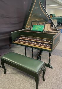 Image of a green harpsichord with open lid and music stand behind a green bench. The case and lid are accented with gold boxes.