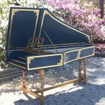 Photograph of a blue harpsichord with gold decorative paint and turned legs. The photo is outdoors and there are azalea hedges in the background