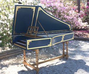 Photograph of a blue harpsichord with gold decorative paint and turned legs. The photo is outdoors and there are azalea hedges in the background
